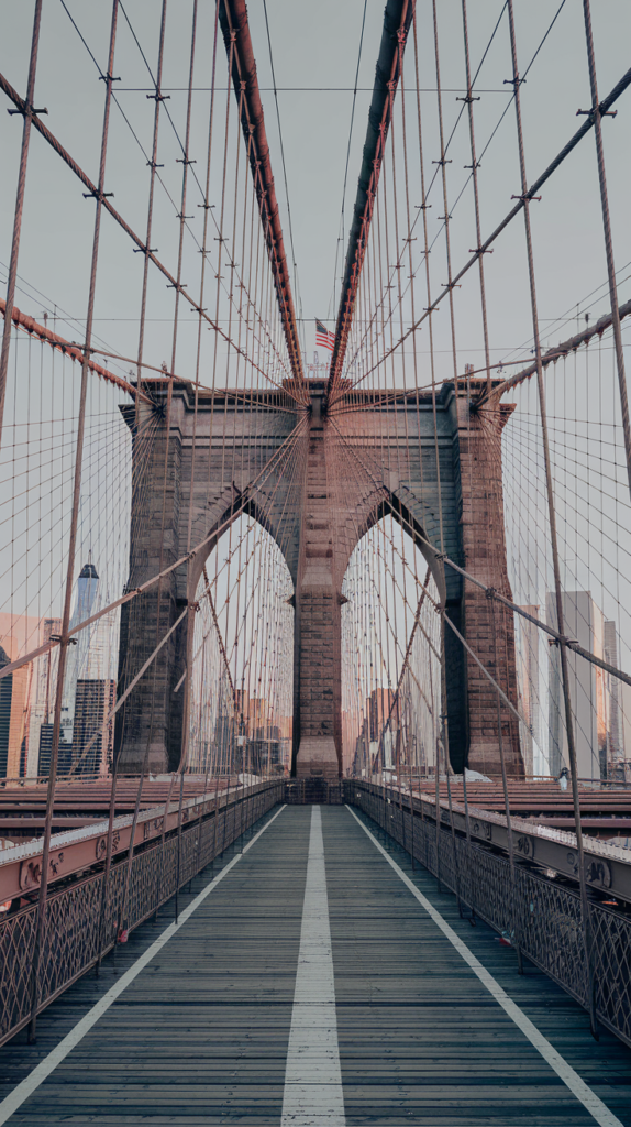 A photo of the Brooklyn Bridge in New York City, taken from the pedestrian walkway. The perspective emphasizes the symmetry of the bridge's suspension cables, which create a striking visual pattern leading toward the stone arches at its center. The city skyline, including notable skyscrapers like One World Trade Center, is visible in the background, bathed in warm, golden-hour light. The wooden walkway and the steel structure of the bridge are detailed and highlight its historic architectural beauty. An American flag flies atop one of the bridge's towers, adding a touch of patriotism to the scene.